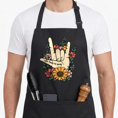 Sign education apron - Beige ASL love hand with Love Needs No Words text, yellow sunflowers, red roses, green leaves — shown on a black cotton canvas apron against a white background