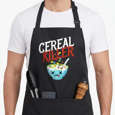 Killer halloween apron - CEREAL KILLER text above blue bowl with dead face and falling colorful loops — shown on a black cotton canvas apron against a white background