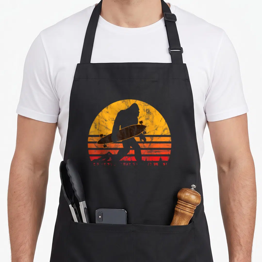 Longboard bigfoot apron - Black gorilla silhouette holding skateboard against striped yellow orange red retro sun — shown on a black cotton canvas apron against a white background