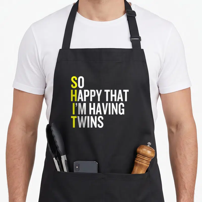 Having gemini girl apron - Stacked text with yellow vertical initials forming an acronym on white background — shown on a black cotton canvas apron against a white background