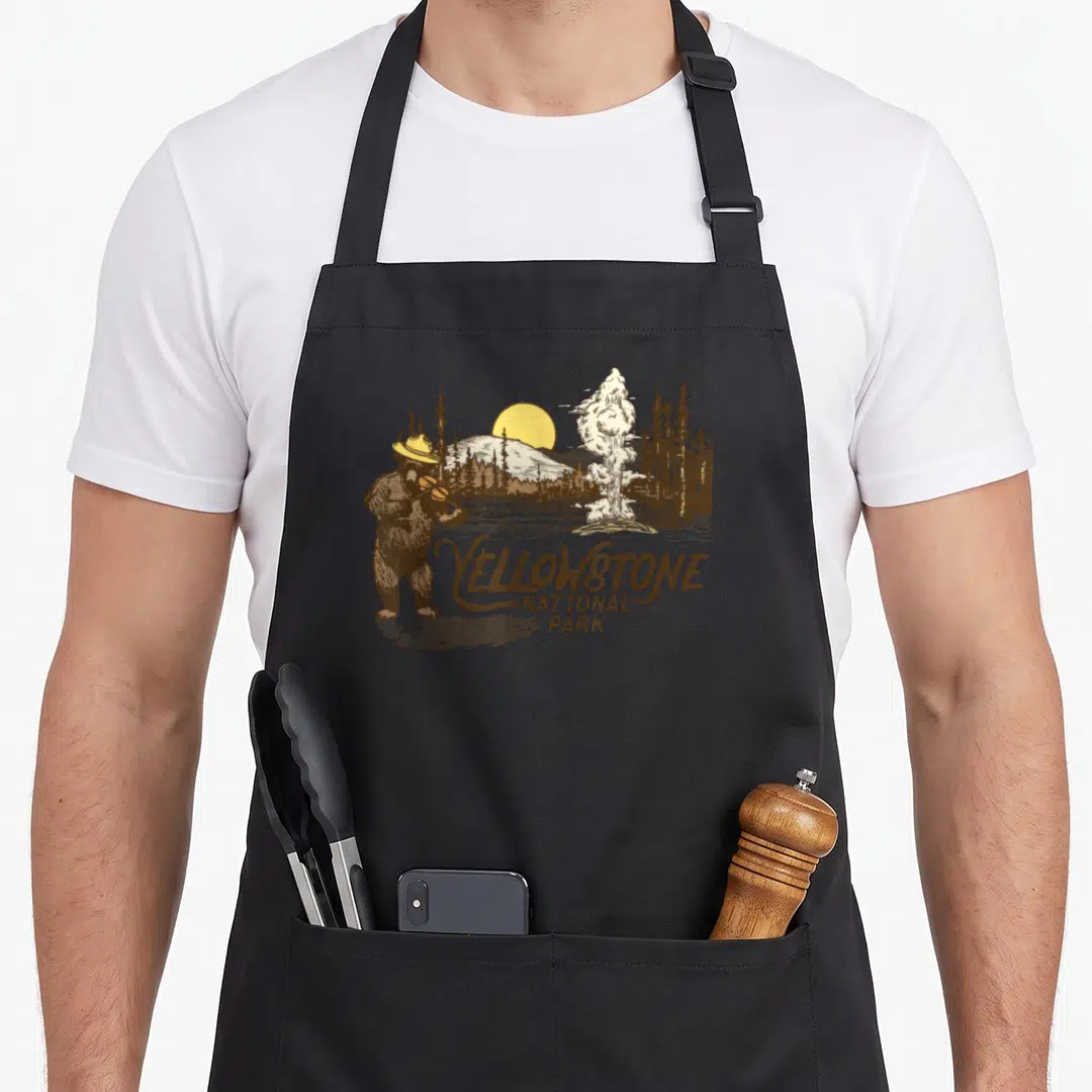 Fiddle bear apron - Brown bear playing violin near white geyser and yellow sun with Yellowstone National Park text — shown on a black cotton canvas apron against a white background