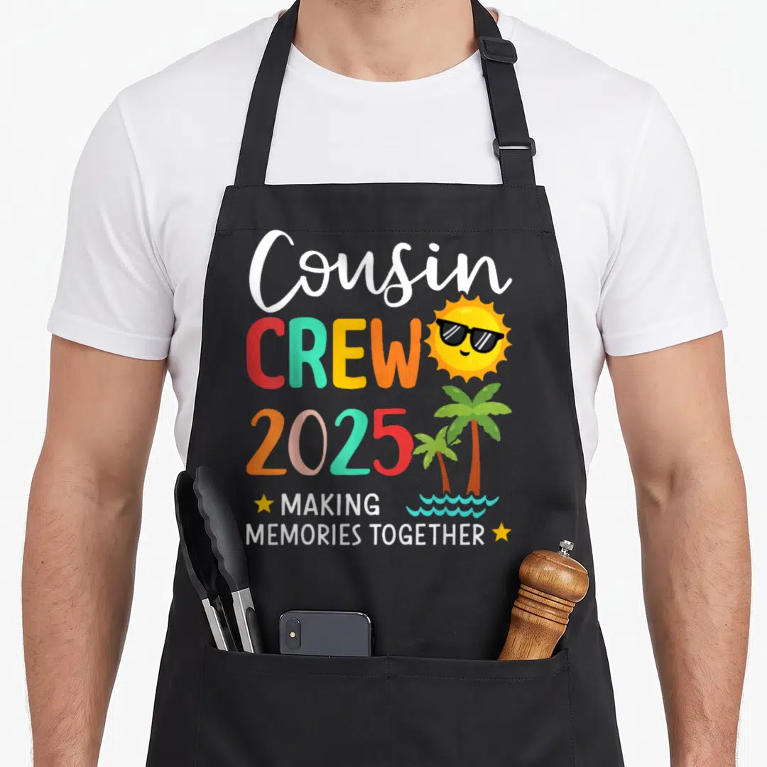 Summer memory apron - Cousin CREW 2025 text with cartoon sun, sunglasses, palm trees, and waves in red teal yellow — shown on a black cotton canvas apron against a white background