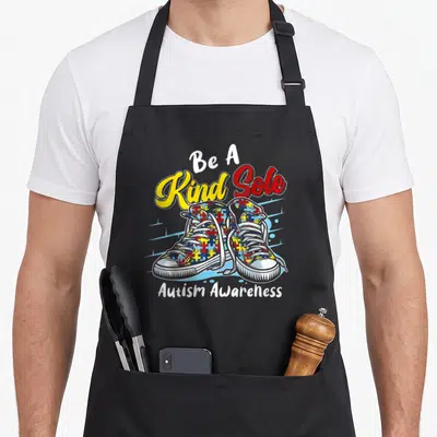 Kind shoe apron - Cartoon sneakers with puzzle pieces and Be A Kind Sole Autism Awareness text — shown on a black cotton canvas apron against a white background