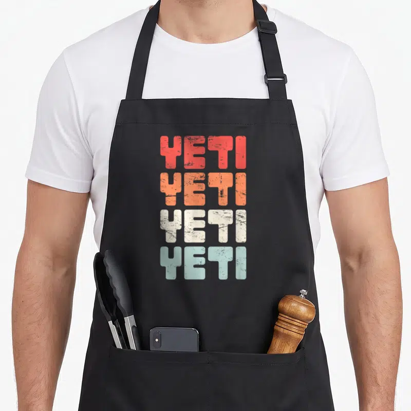 Distressed yeti apron - Stacked YETI text in red orange white and blue distressed block letters — shown on a black cotton canvas apron against a white background