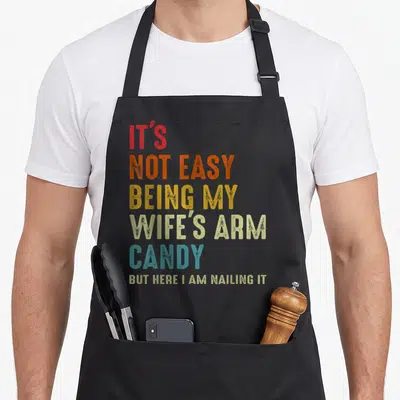 Easy cotton candy apron - Stacked colorful text reading IT'S NOT EASY BEING MY WIFE'S ARM CANDY BUT HERE I AM NAILING IT — shown on a black cotton canvas apron against a white background