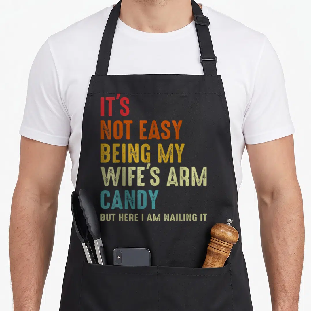 Easy cotton candy apron - Stacked colorful text reading IT'S NOT EASY BEING MY WIFE'S ARM CANDY BUT HERE I AM NAILING IT — shown on a black cotton canvas apron against a white background