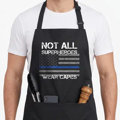 Hero police apron - Distressed text Not All Superheroes Wear Capes over blue line flag — shown on a black cotton canvas apron against a white background