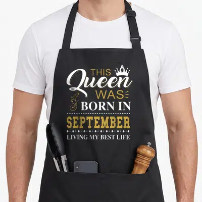 Vintage september baking queen apron - White and gold text reading This Queen Was Born In September Living My Best Life — shown on a black cotton canvas apron against a white background