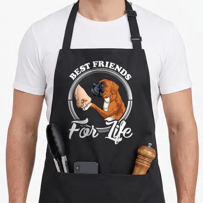Cute boxer dog apron - Cartoon boxer dog fist-bumping human hand with best friends for life text in white circle — shown on a black cotton canvas apron against a white background