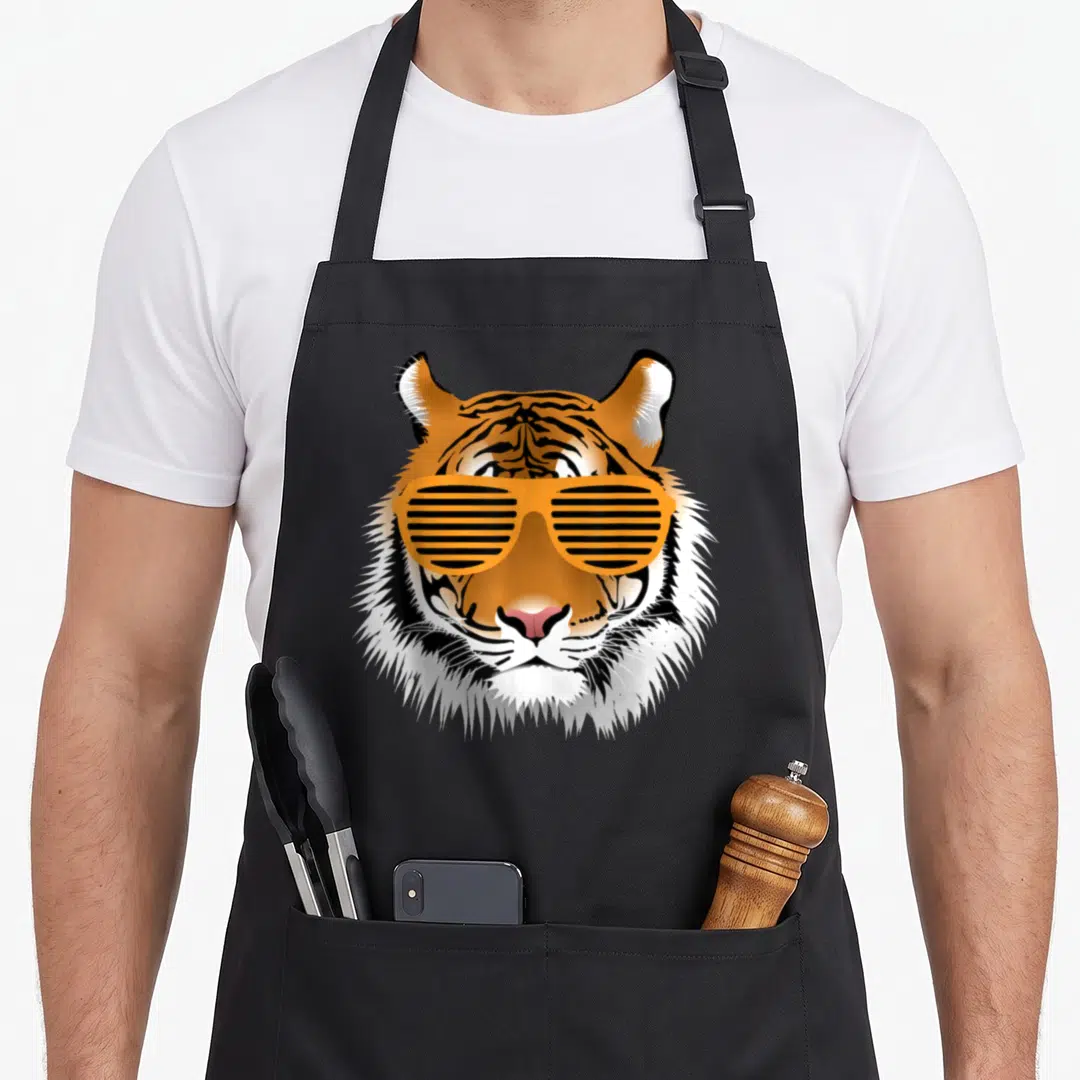 Birthday striped apron - Cartoon tiger head with orange shutter shades on black background — shown on a black cotton canvas apron against a white background