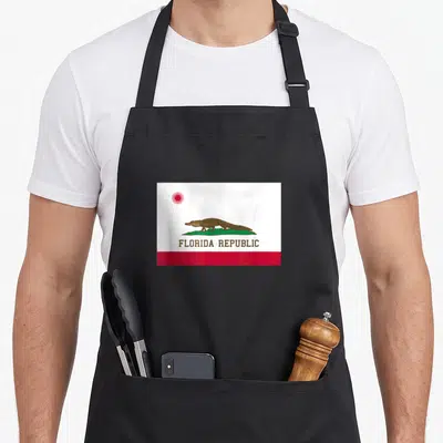 Republic florida apron - Brown alligator on green grass below red sun and FLORIDA REPUBLIC text above red stripe — shown on a black cotton canvas apron against a white background