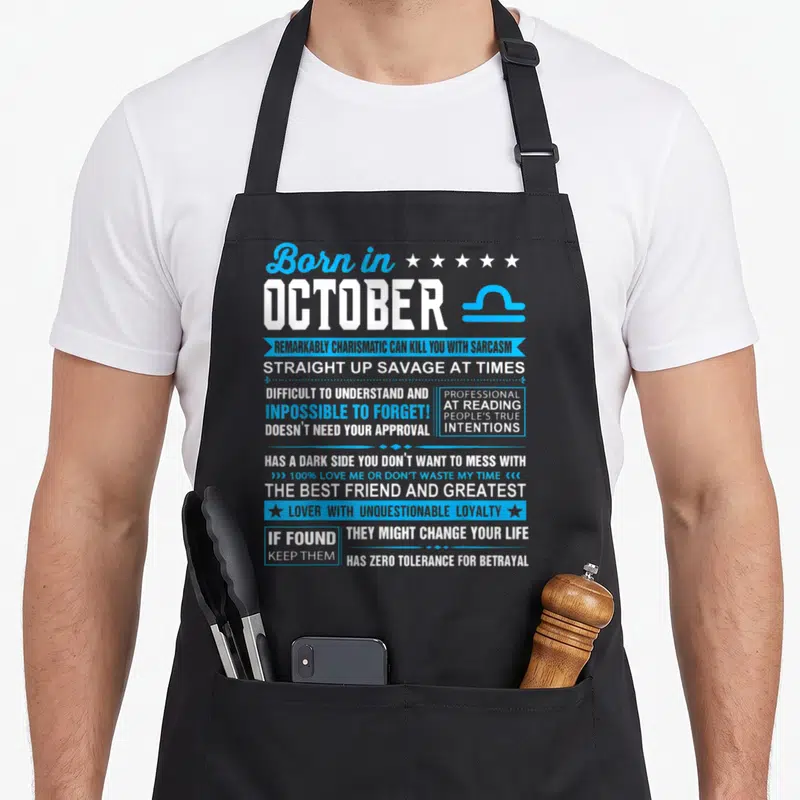 Funny libra birthday apron - Born in October Libra text with blue ribbons, stars, and zodiac symbol listing sarcastic — shown on a black cotton canvas apron against a white background