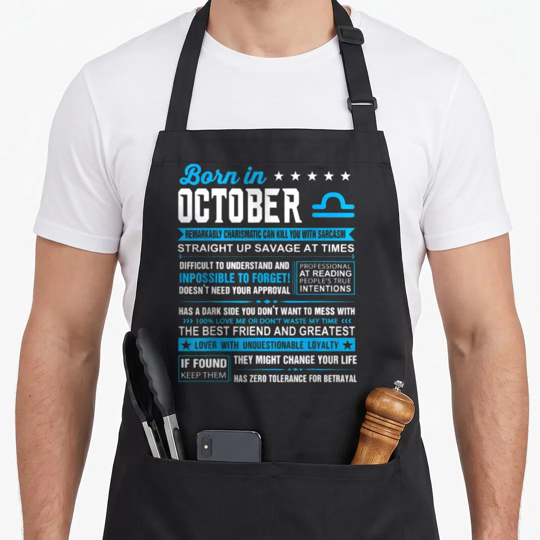 Funny libra birthday apron - Born in October Libra text with blue ribbons, stars, and zodiac symbol listing sarcastic — shown on a black cotton canvas apron against a white background