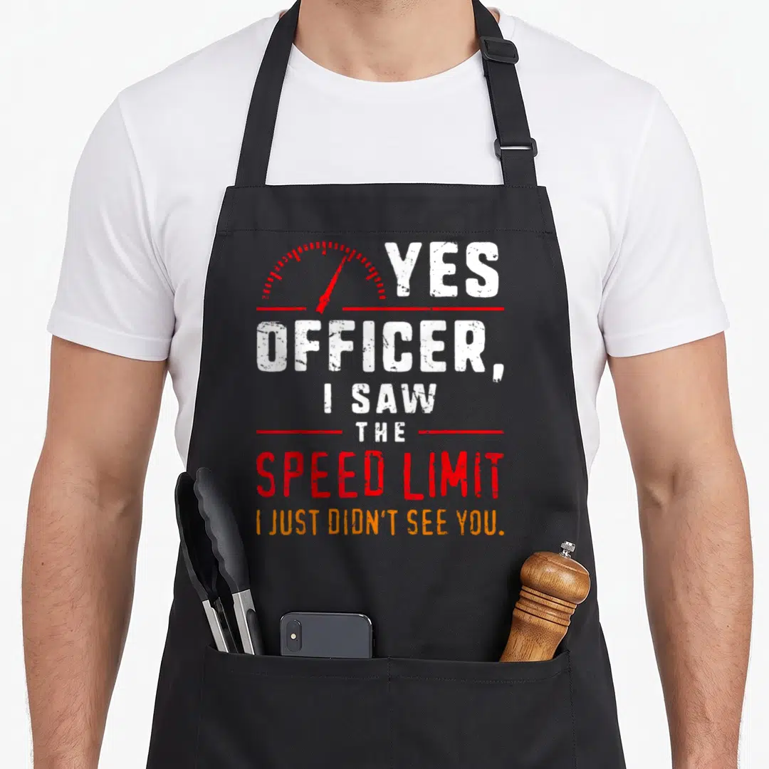 Speeding mechanic apron - Red speedometer arc above stacked white and orange text reading Yes Officer I Saw The Speed — shown on a black cotton canvas apron against a white background