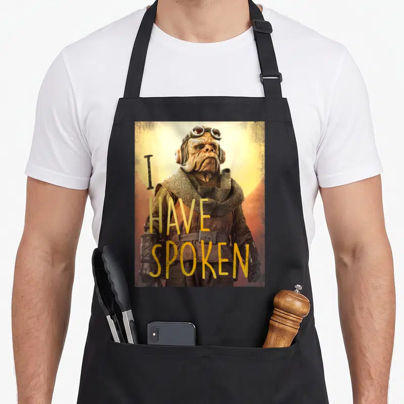 Kuiil i love tribute apron - Brown alien wearing goggles with yellow I HAVE SPOKEN text overlay on circular background — shown on a black cotton canvas apron against a white background