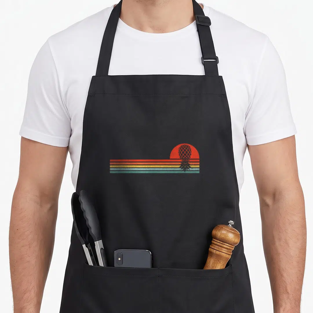 Horizon pineapple apron - Inverted pineapple silhouette inside red sun over orange, yellow, and blue stripes — shown on a black cotton canvas apron against a white background