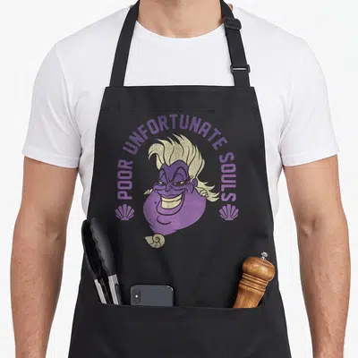 Poor mermaid apron - Ursula cartoon face with POOR UNFORTUNATE SOULS text and two seashells in purple and cream — shown on a black cotton canvas apron against a white background