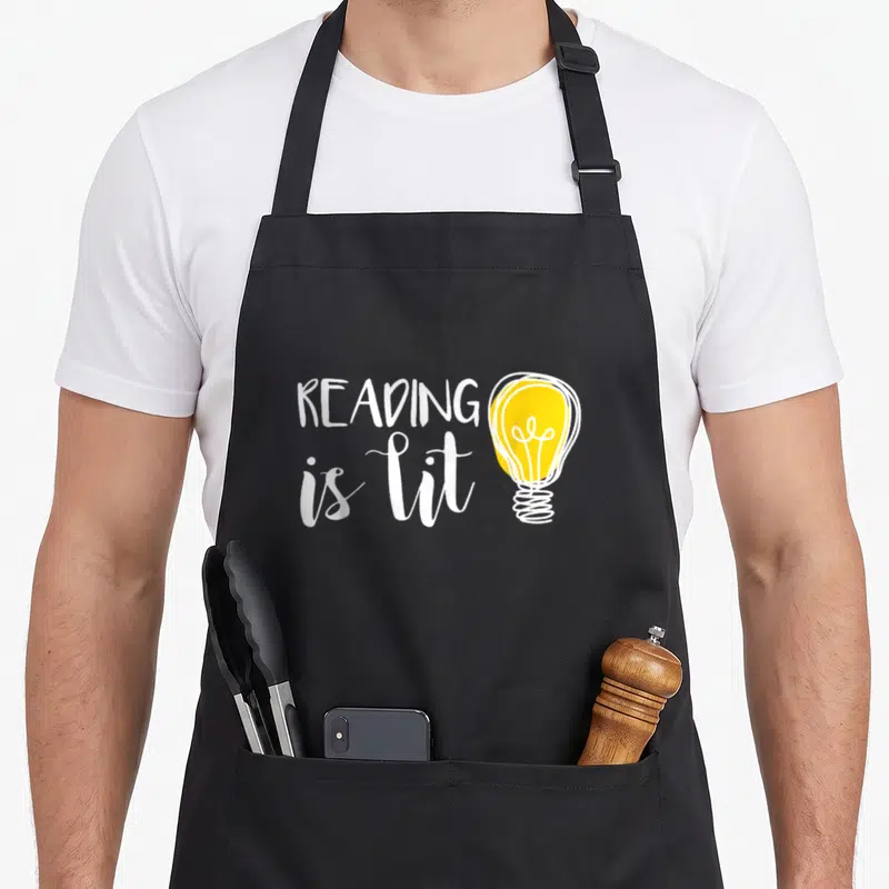 Bookworm teacher apron - White reading is lit text next to yellow cartoon light bulb — shown on a black cotton canvas apron against a white background
