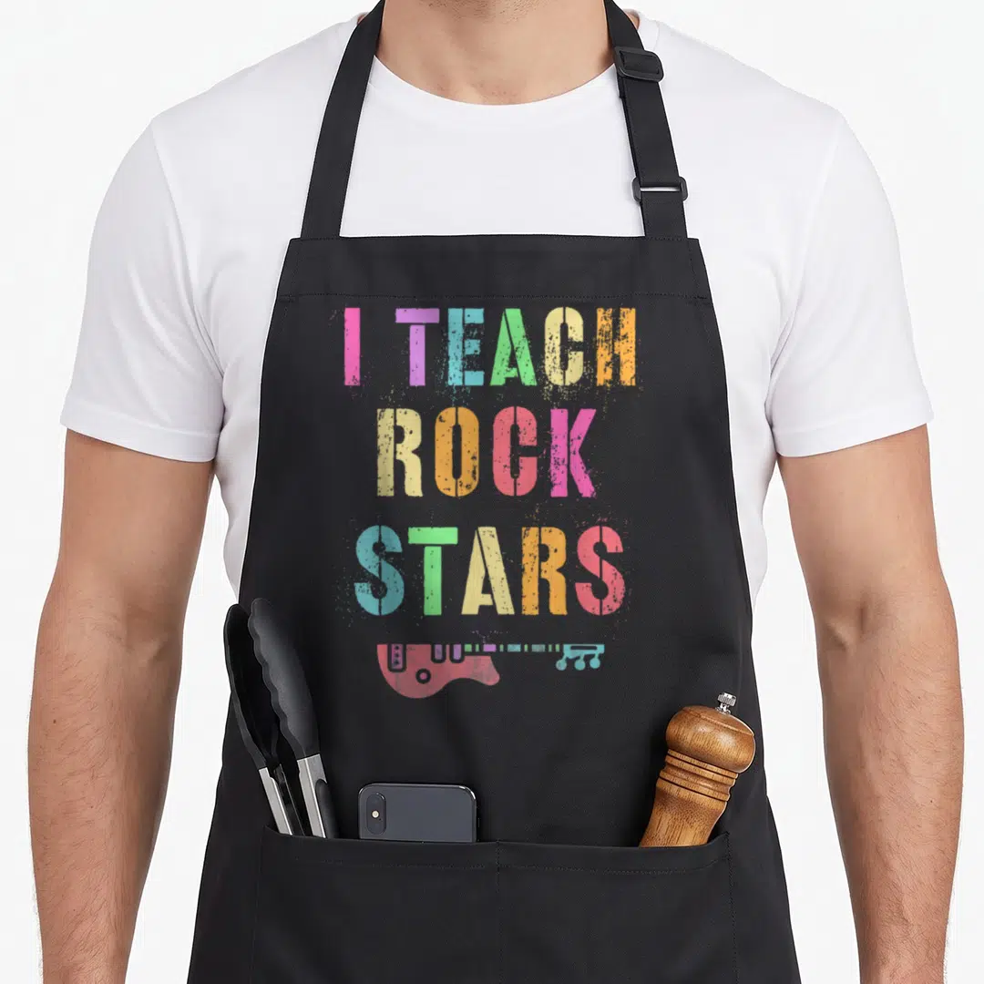Star teacher apron - I TEACH ROCK STARS text in rainbow colors above pink electric guitar with paint splatters — shown on a black cotton canvas apron against a white background