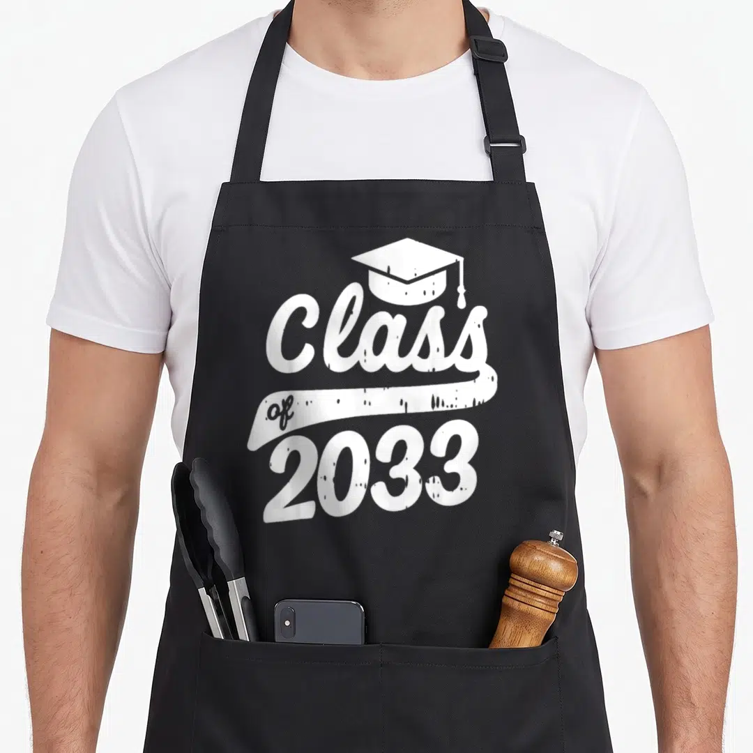 Kindergarten future class apron - White distressed Class of 2033 text with graduation cap and ribbon banner — shown on a black cotton canvas apron against a white background