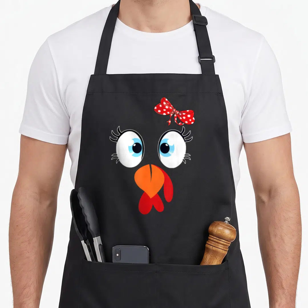 Family thanksgiving aprons - Cartoon chicken face with red polka-dot bow, blue eyes, orange beak, red wattle — shown on a black cotton canvas apron against a white background