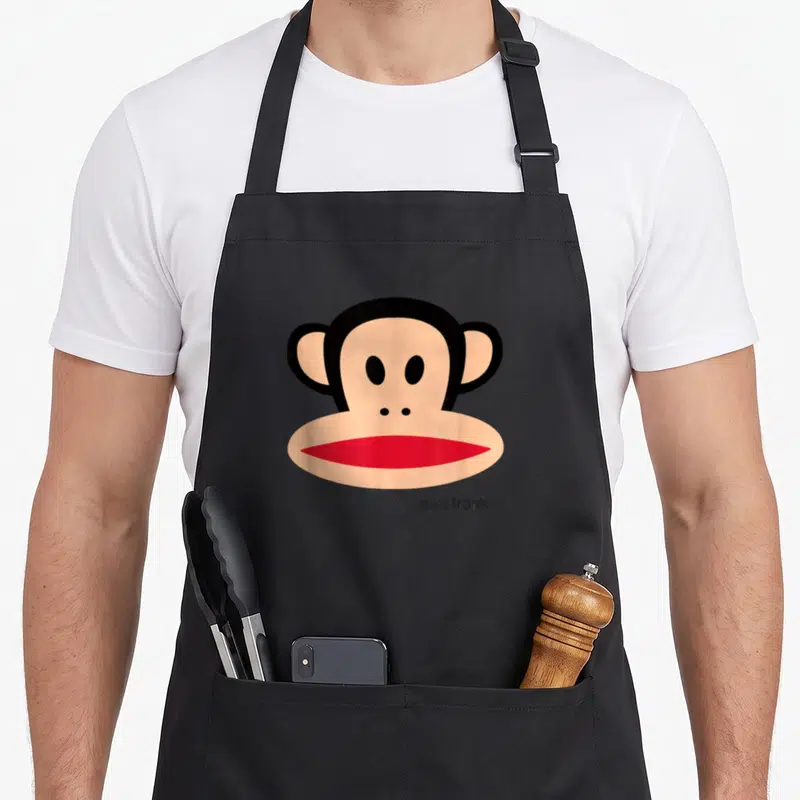 Frank face apron - Tan and black monkey head with red mouth and small logo text at bottom right — shown on a black cotton canvas apron against a white background