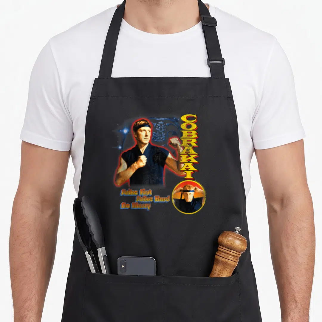 Cobra 90s apron - Realistic martial artist with blue cobra, vertical COBRA KAI text, and three slogan lines — shown on a black cotton canvas apron against a white background