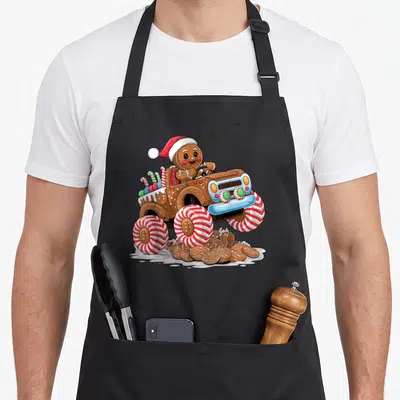 Monster christmas gingerbread apron - Gingerbread man in Santa hat drives monster truck with peppermint tires crushing — shown on a black cotton canvas apron against a white background