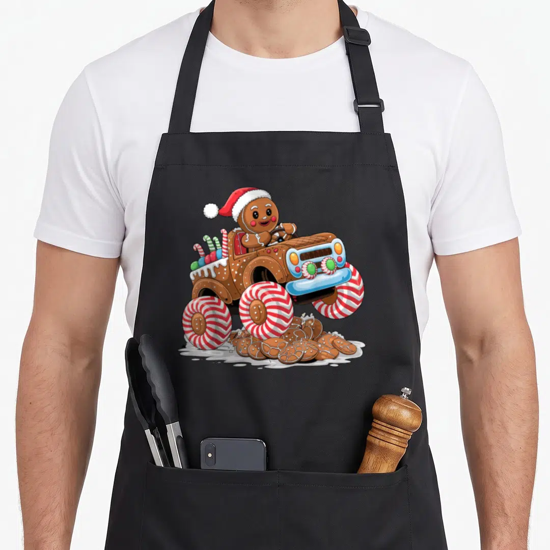 Monster christmas gingerbread apron - Gingerbread man in Santa hat drives monster truck with peppermint tires crushing — shown on a black cotton canvas apron against a white background