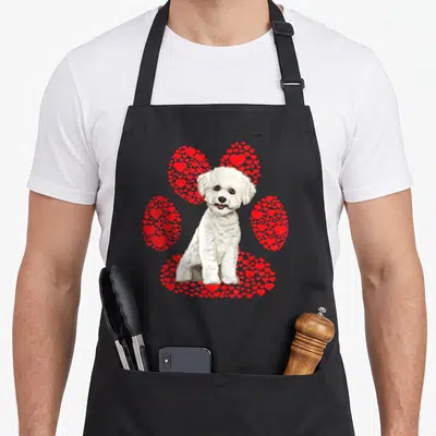 Bichon valentine's day apron - Realistic white dog sitting in front of a paw print formed by red hearts — shown on a black cotton canvas apron against a white background