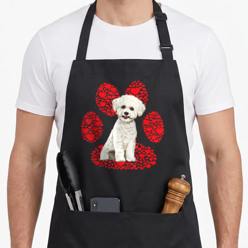 Bichon valentine's day apron - Realistic white dog sitting in front of a paw print formed by red hearts — shown on a black cotton canvas apron against a white background