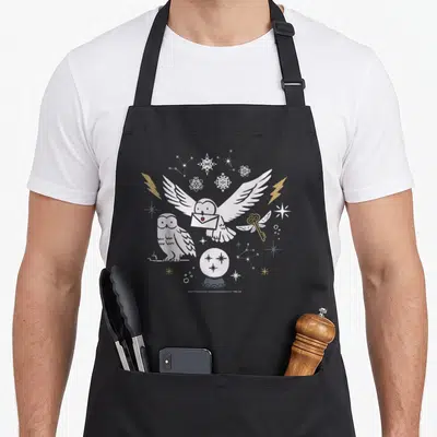 Owl winter apron - Flying owl holding letter, crystal ball, golden key, snowflakes, and lightning bolts in white gold — shown on a black cotton canvas apron against a white background