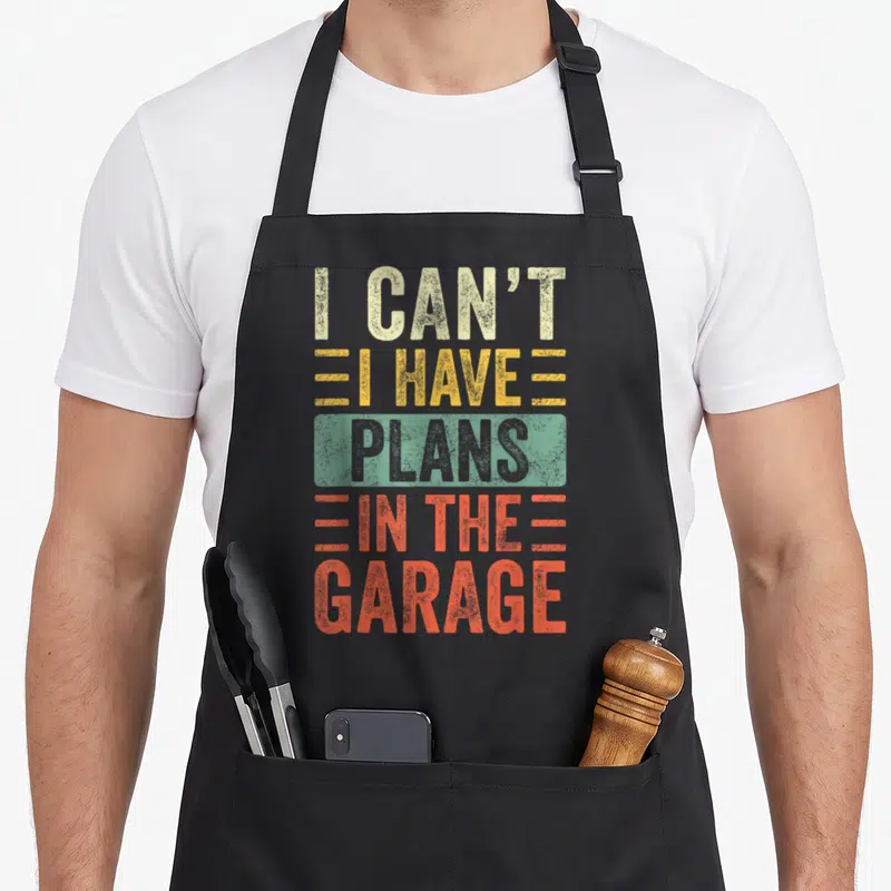 Plan car garage apron - Stacked distressed text reading I CAN'T I HAVE PLANS IN THE GARAGE in beige yellow grey — shown on a black cotton canvas apron against a white background