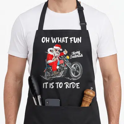 Biker merry christmas apron - Santa in red suit riding motorcycle with Oh What Fun It Is To Ride text — shown on a black cotton canvas apron against a white background