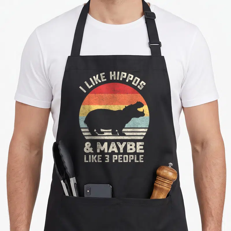 Funny hippo animal apron - Retro striped sun with hippo silhouette and text I like hippos maybe like 3 people — shown on a black cotton canvas apron against a white background