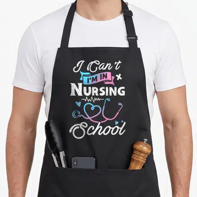 Funny school nursing apron - I Can't I'm In Nursing School text with gradient stethoscope heart and EKG line — shown on a black cotton canvas apron against a white background