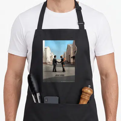 Wish pink floyd apron - Two men in suits shaking hands, one engulfed in orange flames between industrial buildings — shown on a black cotton canvas apron against a white background