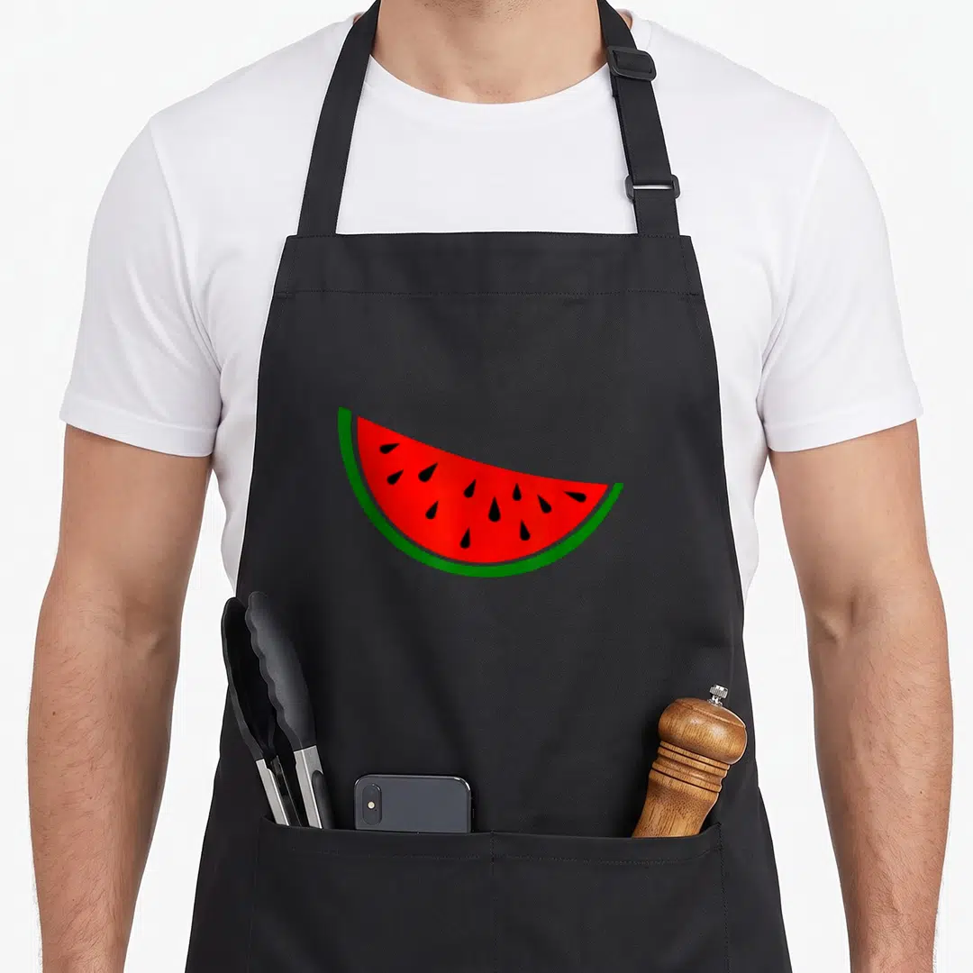 Funny watermelon apron - Cartoon watermelon slice with red flesh green rind and black seeds — shown on a black cotton canvas apron against a white background