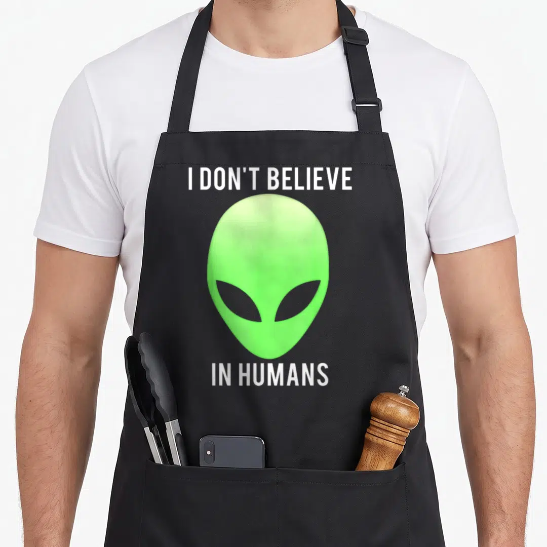 Believe alien apron - Lime green alien head between white text reading I DON'T BELIEVE IN HUMANS — shown on a black cotton canvas apron against a white background