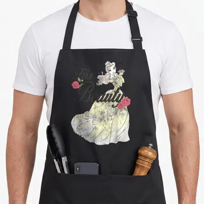 Belle bridal apron - His Beauty text over cartoon woman in yellow dress flanked by red roses on grey background — shown on a black cotton canvas apron against a white background