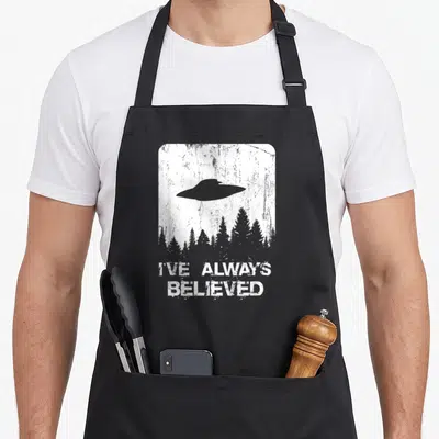 Abduction alien ufo apron - Grey UFO over forest trees with I've Always Believed text on distressed white background — shown on a black cotton canvas apron against a white background