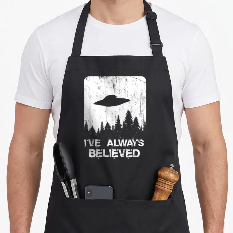 Abduction alien ufo apron - Grey UFO over forest trees with I've Always Believed text on distressed white background — shown on a black cotton canvas apron against a white background