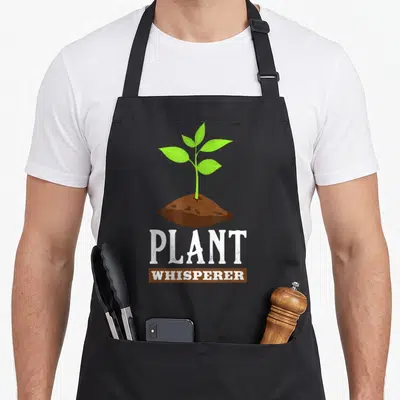 Landscaper gardener apron - Green sprout rising from brown soil mound above white Plant Whisperer text — shown on a black cotton canvas apron against a white background