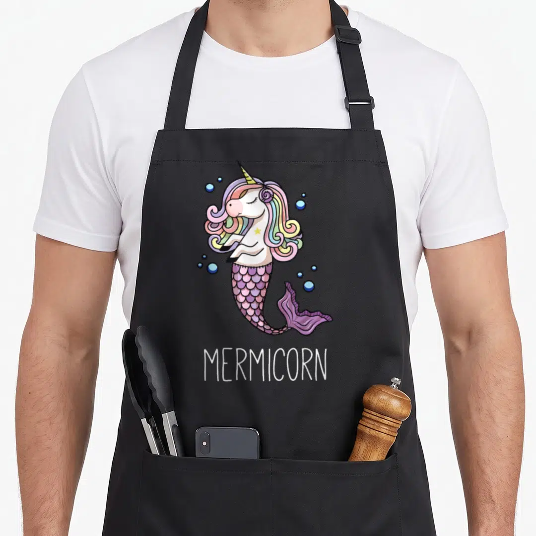 Mermicorn unicorn apron - Cartoon mermicorn with rainbow mane, purple tail, blue bubbles, and centered white text — shown on a black cotton canvas apron against a white background