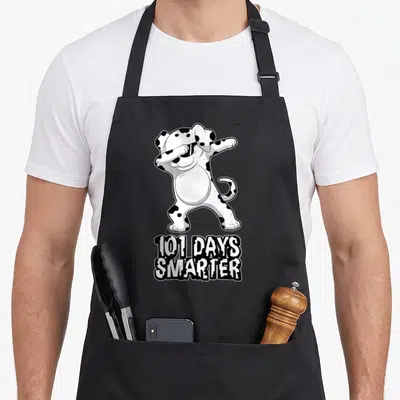 Teacher dog apron - Cartoon dalmatian dabbing above 101 Days Smarter text in black and white — shown on a black cotton canvas apron against a white background