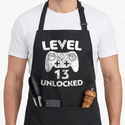 Gamer 13th birthday apron - White distressed game controller between LEVEL 13 UNLOCKED text on black apron — shown on a black cotton canvas apron against a white background