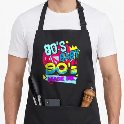 Baby 90s 80s apron - Stacked 80S BABY 90S MADE ME text in magenta yellow and blue with geometric shapes — shown on a black cotton canvas apron against a white background