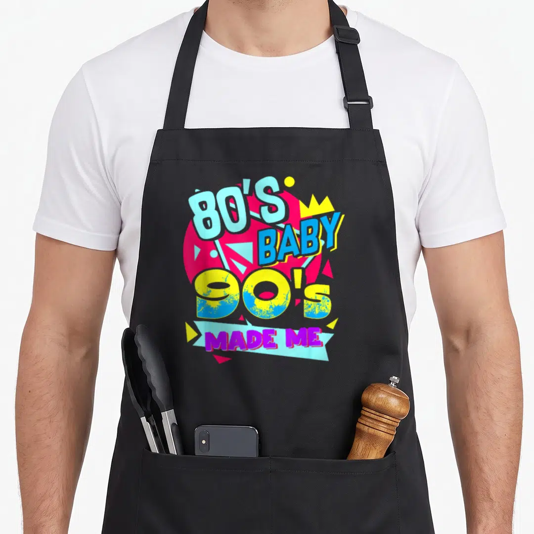 Baby 90s 80s apron - Stacked 80S BABY 90S MADE ME text in magenta yellow and blue with geometric shapes — shown on a black cotton canvas apron against a white background