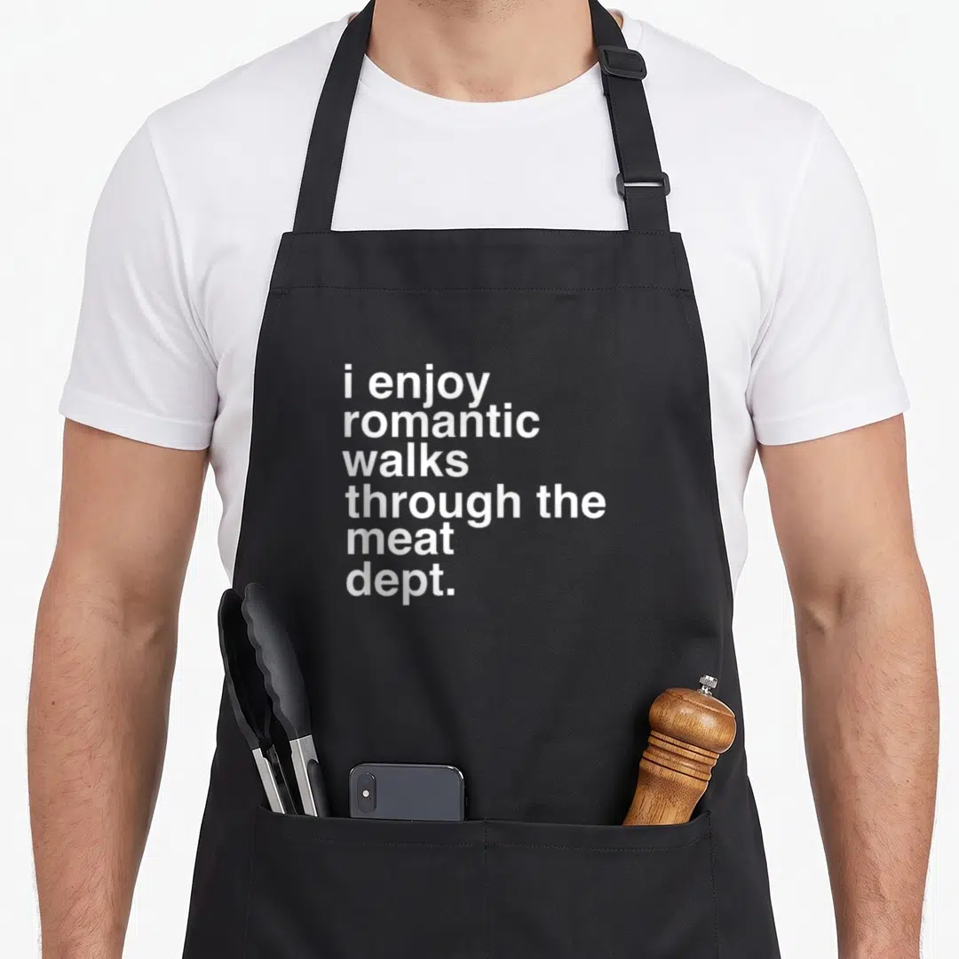 Bacon meat aprons - White text i enjoy romantic walks through the meat dept on black background — shown on a black cotton canvas apron against a white background