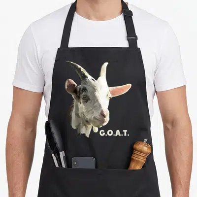Cute greatest apron - Realistic goat head illustration with G.O.A.T. text in white, grey, and brown tones — shown on a black cotton canvas apron against a white background
